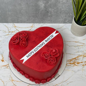 Heart-shaped red cake with rose decorations and a banner reading Happy Birthday, March on a marble surface next to a potted plant.