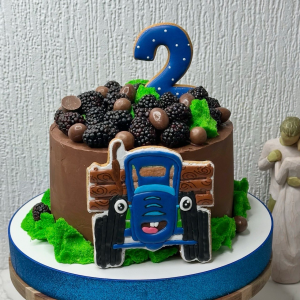 Chocolate cake topped with blackberries, chocolate sweets, a blue number 2 candle, green icing, and a blue lorry biscuit; figurine in background.
