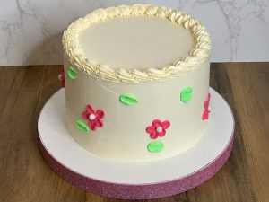 A round cake with white icing, pink and green flower decorations, and a piped border, displayed on a white and pink cake board.