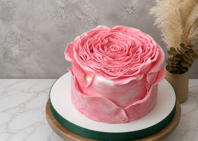 A round cake decorated with pink, petal-like icing to resemble a large flower, placed on a white and green cake board.